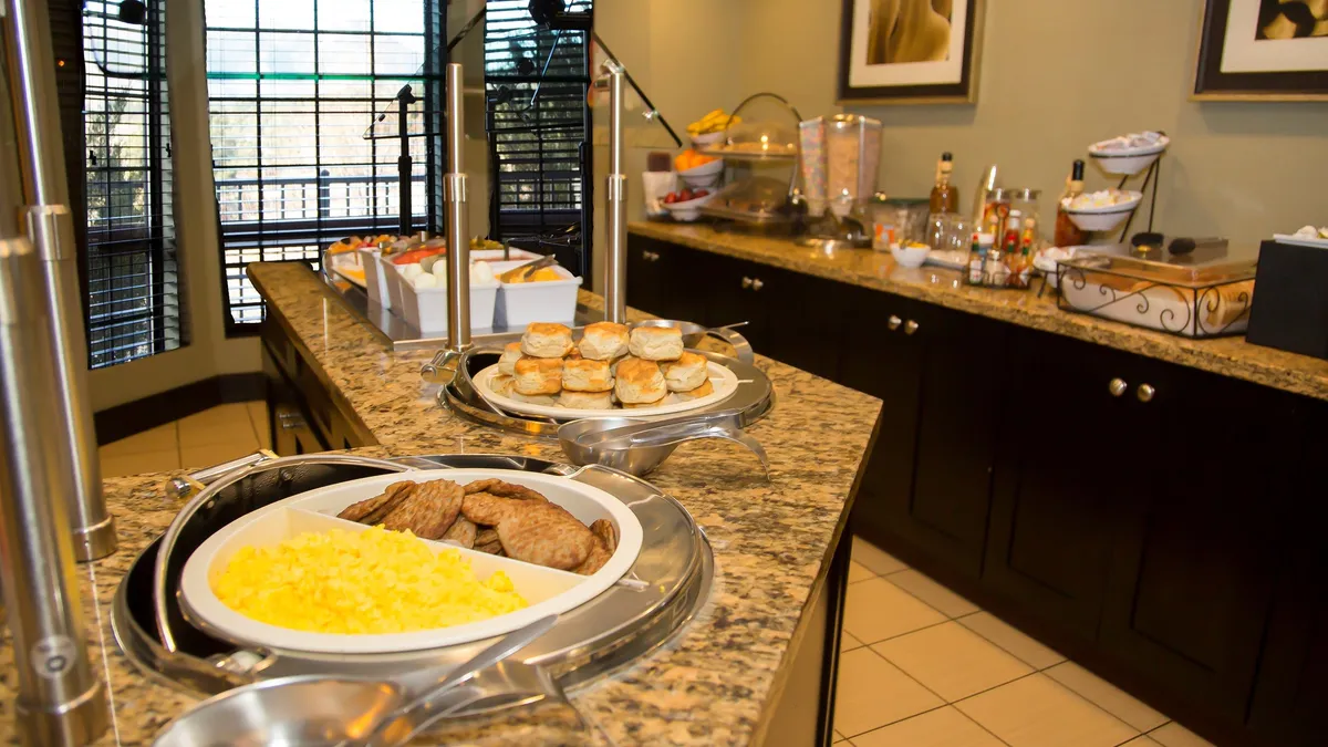 A warmly lit kitchen scene featuring a granite countertop set with a fresh food display and a welcoming coffee station. The background hints at a communal atmosphere, with enough space for both cooking and gathering. Essential appliances and kitchen amenities are subtly visible, supporting the narrative of a fully equipped culinary area.
