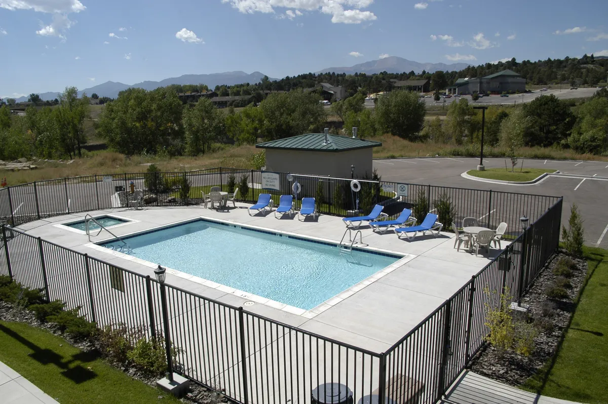 A bright exterior shot showcasing the swimming pool area with lounge chairs and a fenced perimeter, set against a backdrop of scenic landscape. The vibe is leisurely, hinting at lazy afternoons spent relaxing, sunbathing, or socializing by the pool.