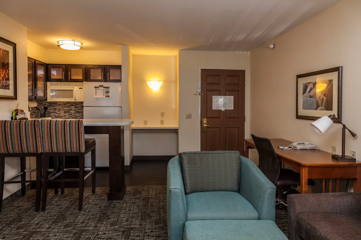This image showcases a bright, inviting studio space with a modern kitchenette, two bar stools ideal for casual meals, a cozy sofa for relaxing, and a functional desk for work or planning adventures. The layout is open and thoughtfully designed, making it perfect for both downtime and productivity.